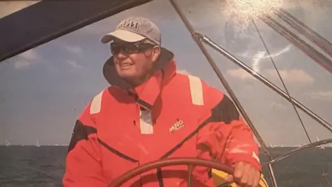 Victor Tinley is holding the wheel of a boat, looking off into the distance with the sea behind him. He is wearing a red waterproof and a grey cap and sunglasses. He is smiling.