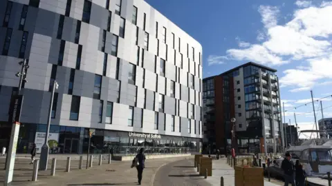 The exterior of the University of Suffolk at Ipswich Waterfront. It is a tall grey-cladded building with many windows. Behind it is a modern block of flats, and to the right is a yacht parked up in the quay.
