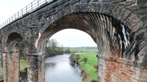 National Highways A close-up view of a viaduct looking through one semicircular stone arch to the river beyond. The odd diagonal stonework of the arch is clearly visible. It spans a river which can be seen in the background, with green fields to either side.