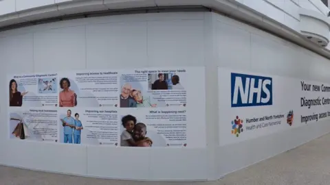 A photo of the exterior of a community diagnostic centre. It shows white walls and banners with photos of people, writing and a large NHS logo. 