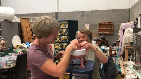 BBC / Naj Modak Sarah Wintringham is pictured in a purple T-shirt showing Ann Ruddick how to use a knitting device