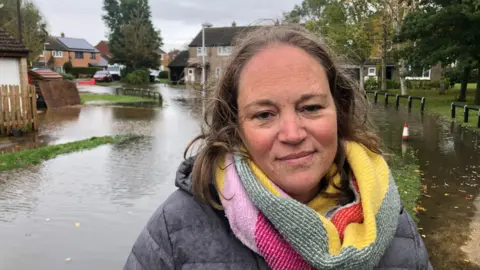 John Devine/BBC Vhari Russell has shoulder length brown hair, a grey coat with colourful woollen scarf and is standing in front of a flooded housing estate.