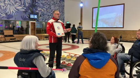 Coach Rick Hills discusses tactics with members of the Preston Wheelchair Curling Club. They are in their wheelchairs on the ice.