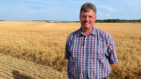 Chris Swanton is standing in a wheat field wearing a checked shirt. He is smiling.
