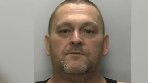 Devon & Cornwall Police A man, with a small beard and brown eyes, looking directly at the camera. There is a grey wall behind him. 