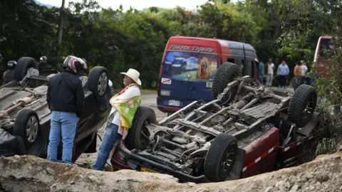 Two people in a motorcycle helmet and white hat look on while surverying the scene which includes two upside down vehicles in front of a bus parked on the side of the road.