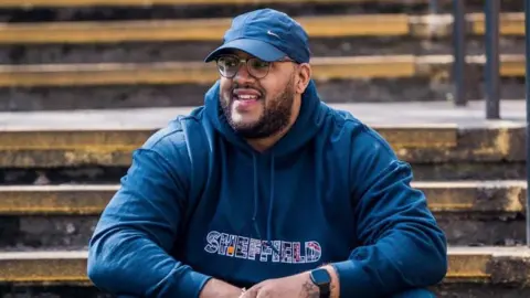 Simon Thake/BBC A man in his 30s or 40s wearing a navy hoodie with "Sheffield" written on it in capital letters. He has a navy cap on with a white Nike tick on the front. He has glasses and a black beard and is looking off camera, smiling. He appears to be sat on some outdoor stairs.