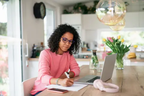 Getty Images A woman sits at her kitchen table with a laptop and pen and paper