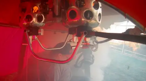 AAIB A person is inside a hot air balloon which has a red envelope. There is still fire alight inside the burners. There is smoke coming from the hot air balloon which is landing in a field.