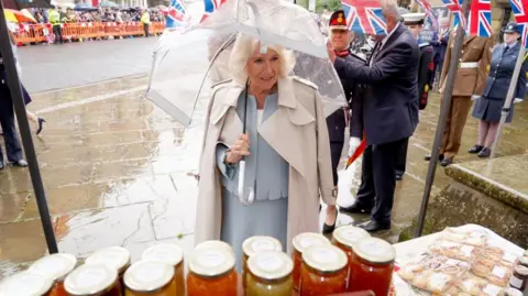 PA The Queen in a grey suit holding a clear plastic umbrella with a rain coat draped over her shoulders. There are crowds behind barriers behind her. There are jars and baked goods on a table in front of her. A man and woman in military uniform are standing behind her.