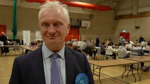 MP Graham Stuart smiles at the election count. He is wearing a Conservative Party rosette. 