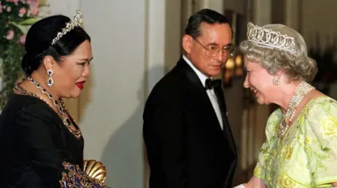 Reuters Queen Sirikit, wearing a black dress, greets Queen Elizabeth II, wearing a light freen dress and white gloves. Thailand's late King Bhumibol stands in the background between them.