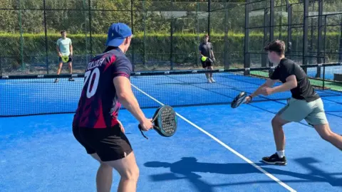Charlie Stubbs / BBC There are four men playing padel. On the one side, one of the men has a lime green t-shirt and black shorts with a yellow racquet. The other man is wearing a black t-shirt and black shorts. In the front, there is a man with his back to camera wearing a blue backwards cap, a purple, pink and navy shirt with 'Tulloch 70' on the back and black shorts. He is holding a black raquet. On the right, at the front, there is a man with a black t-shirt, cyan shorts and black shoes. He is hitting the ball.