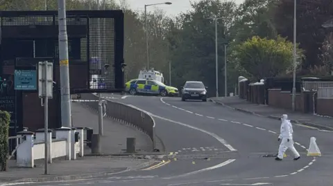 Aity street outside a police station is blocked by police. A person wearing a white forensic suit is walking across the road. Small evidence markers are scattered on the ground, and a police car is parked farther up the street.
