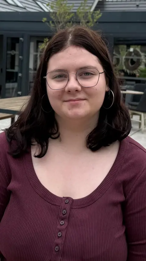 Aoísha is a young girl with shoulder-length dark hair. She is wearing a pair of wire glasses and large silver hoops. She is wearing a burgundy, ribbed shirt with buttons down the middle.