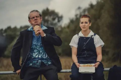 Jessica Sansom Two characters in the film sitting side by side. On the left is an older man who is looking thoughtful while eating. He is wearing a black suit with a Hawaiian-style blue shirt underneath. The younger woman sitting to his right has red hair pulled back into a ponytail. She is wearing a white, short sleeved shirt and blue dungarees. She is holding a box of chips.