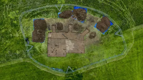 An aerial shot of the dig site. One main area is excavated in a rough square shape, with a series of smaller, rounder dig sites around it. 