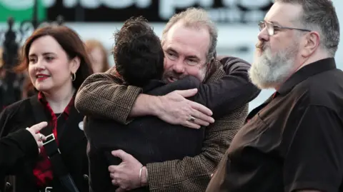 PA Media Gavin Mitchell hugs a antheral   successful  the mediate  of shot, astatine  the close    is simply a ample  antheral   with grey heair and glasses and  a younger pistillate   with reddish  lipstick connected  the left. All are wearing acheronian  colours.