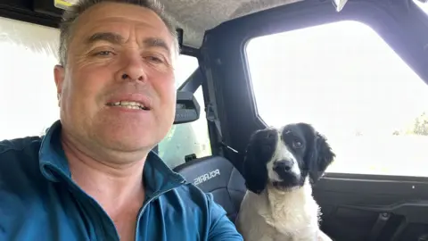 Thomas Conway A man is pictured in the front seat of a jeep, with a springer spaniel dog sitting in the passenger side seat. the man is wearing a blue jumper. the dog is black and white in colour.