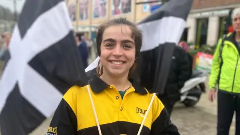 Imagen de una persona con un polo amarillo y negro. Sostiene dos banderas de Cornualles a cada lado.