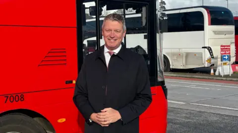 Luke Pollard wearing a black coat and shirt and tie standing smiling in front of a red double decker bus