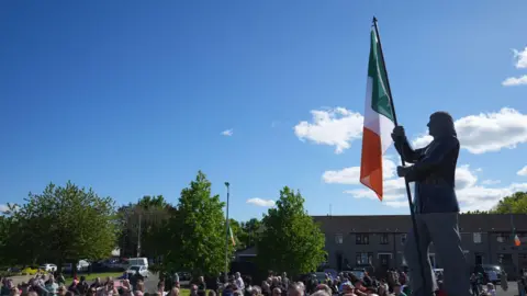 PA Media Statue of Bobby Sands in west Belfast. It is a grey statue and there is an Irish tricolour being held up. There is a crowd nearby.