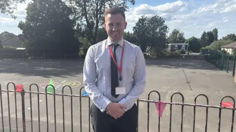 Hill Avenue Academy A man in a shirt stands in a school playground with a tie on and smiling.