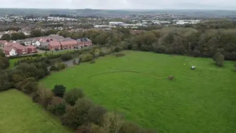 North Devon Council The picture shows a wide stretch of green farmland bordered by trees, with a housing estate sitting along the left edge of the scene. The homes are arranged in neat rows with tiled roofs, and they back directly onto the open field. The field itself is mostly flat.