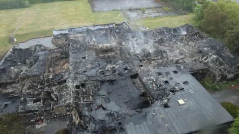 Suffolk Fire and Rescue Service An aerial view of a building that has been destroyed in a fire. The building is covered in ash and soot and some smoke is rising from the site.