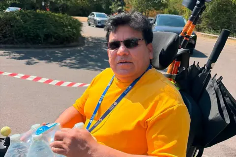 Abdul Jamil, who has dark hair and sunglasses. He is in a wheelchair, wearing an orange top and holding a pack of water bottles.