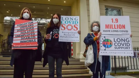 Getty Images Protesters hold placards calling for more research and support for people struggling with Long Covid during the demonstration. Protesters and bereaved family members gathered outside the Covid-19 Inquiry Hearing Centre as former prime minister Boris Johnson gave his testimony on the second day.