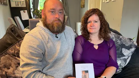 A man and woman sitting next to each other on a sofa in a living room. They are holding an order of service from a funeral