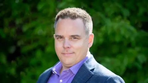 A man looking at the camera wearing a purple shirt and blue jacket. He has short dark, spiky hair and is smiling.  