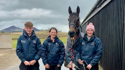 Three people in navy jackets stand beside a racehorse wearing a blanket outside a stable, with rolling hills and a cloudy sky in the background