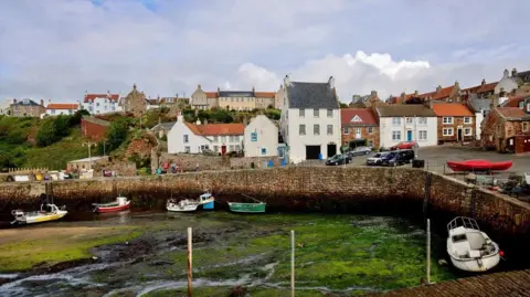 Graham Christie Small fishing boats sit in a harbour at low tide, surrounded by seaweed-covered mud and stone harbour walls. Behind them, a row of white and stone houses with red and grey roofs lines the waterfront under a cloudy sky.