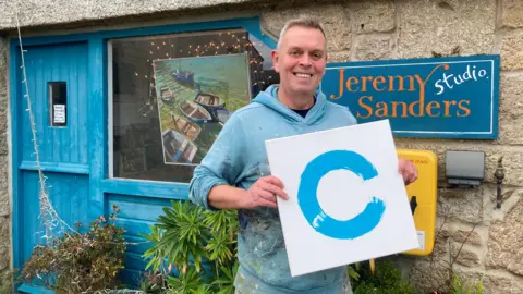 A smiling man in a paint-splattered blue hoodie holds a white canvas with a blue pain-effect on it. He is standing in front of a granite art studio with a weathered blue door, plants and a sign saying Jeremy Sanders Studio.