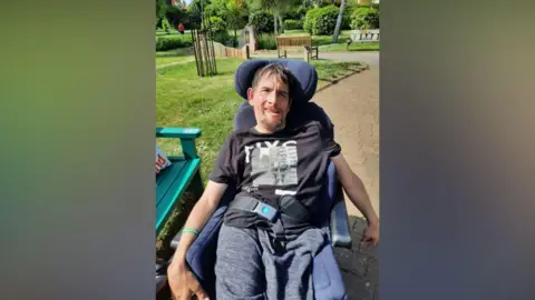 Tom Lewis Tom is sitting outside in the sunshine in a wheelchair next to a green bench in a park. He is smiling to camera