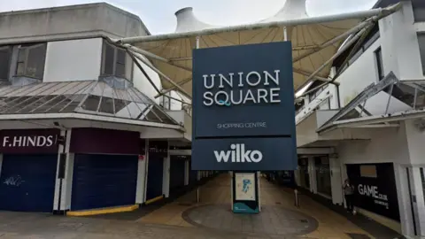 The outside of the Union Square shopping centre in Torquay. There is a large square navy blue sign that reads Union Square with shops either side. 