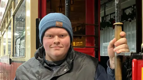 GUY CAMPBELL/BBC Alfie Blanchflower is wearing a grey jacket and blue hat and is standing in the entrance of a red and white tram