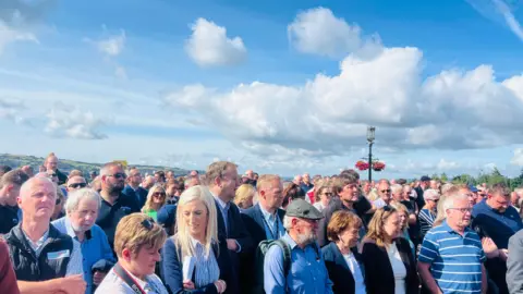 A large crowd of men and women stand facing Stormont on a sunny day.  There are trees and fields in the distance behind them. 