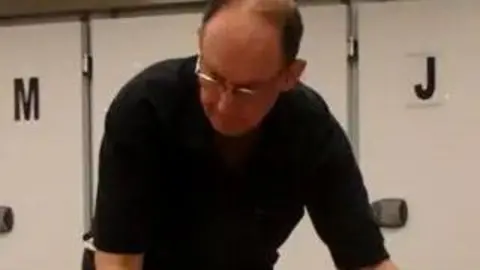 Kent Police A man wearing a black T shirt and glasses is seen leaning forwards towards a video camera. There are mortuary fridges in the background.