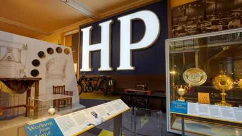 David Rowan/Birmingham Museums Trust The letters HP are lit up on a wall with various other items in cabinets in the museum on display around it