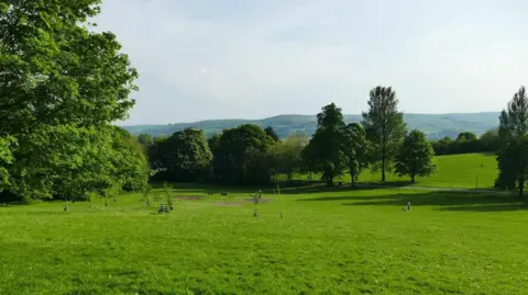 Geograph/Stephen Craven A large green space with several shrubs and trees. Rolling hills can be seen in the distance on what is a sunny day.