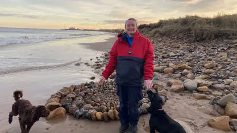 Marina Bullough is standing next to the sculpture. She has two dogs with her and is wearing blue trousers and a red and blue jacket. 