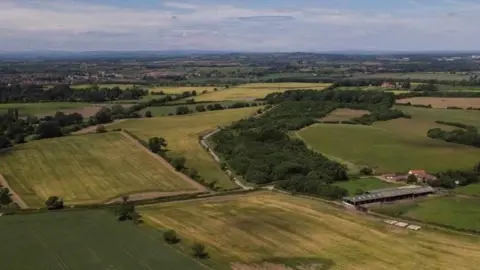 An aerial view of the future site of Skerningham Garden Village in Darlington. It shows a number of green fields and woodland. A farm building can be seen to the right.