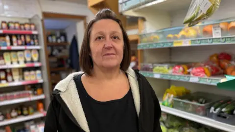 A woman is looking at the camera, she has dark hair in a shoulder-length bob. She is wearing a black t-shirt and a black hoody with a white sheppskin lining. She is stood in a shop with fruit and veg to the right of her and bottled goods on a shelf to the left of her. 