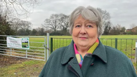 Chris is looking into the camera and smiling slightly. She's wearing a green wool overcoat, with a brightly coloured scarf underneath. Behind her you can see the Glebe, a large green field with trees and bushes around the edges. Between Chris and the field is a large silver padlocked gate, and waist-height green wrought iron fencing.