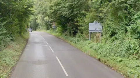 A road surrounded by trees and bushes either side. There is a yellow speed limit sign on the right as well as a white sign which says "Much Wenlock"