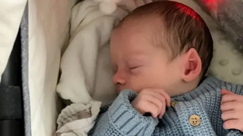 A baby is asleep in his carrycot. He is wearing a pale blue knitted cardigan, his head is turned to the left. He has light brown hair. He is lying on light coloured, cosy blankets.