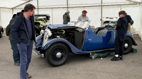 A group of people admire a blue classic car in a marquee. The bonnet has been lifted up to show the engine. A number of other classic cars can be seen in the background. 